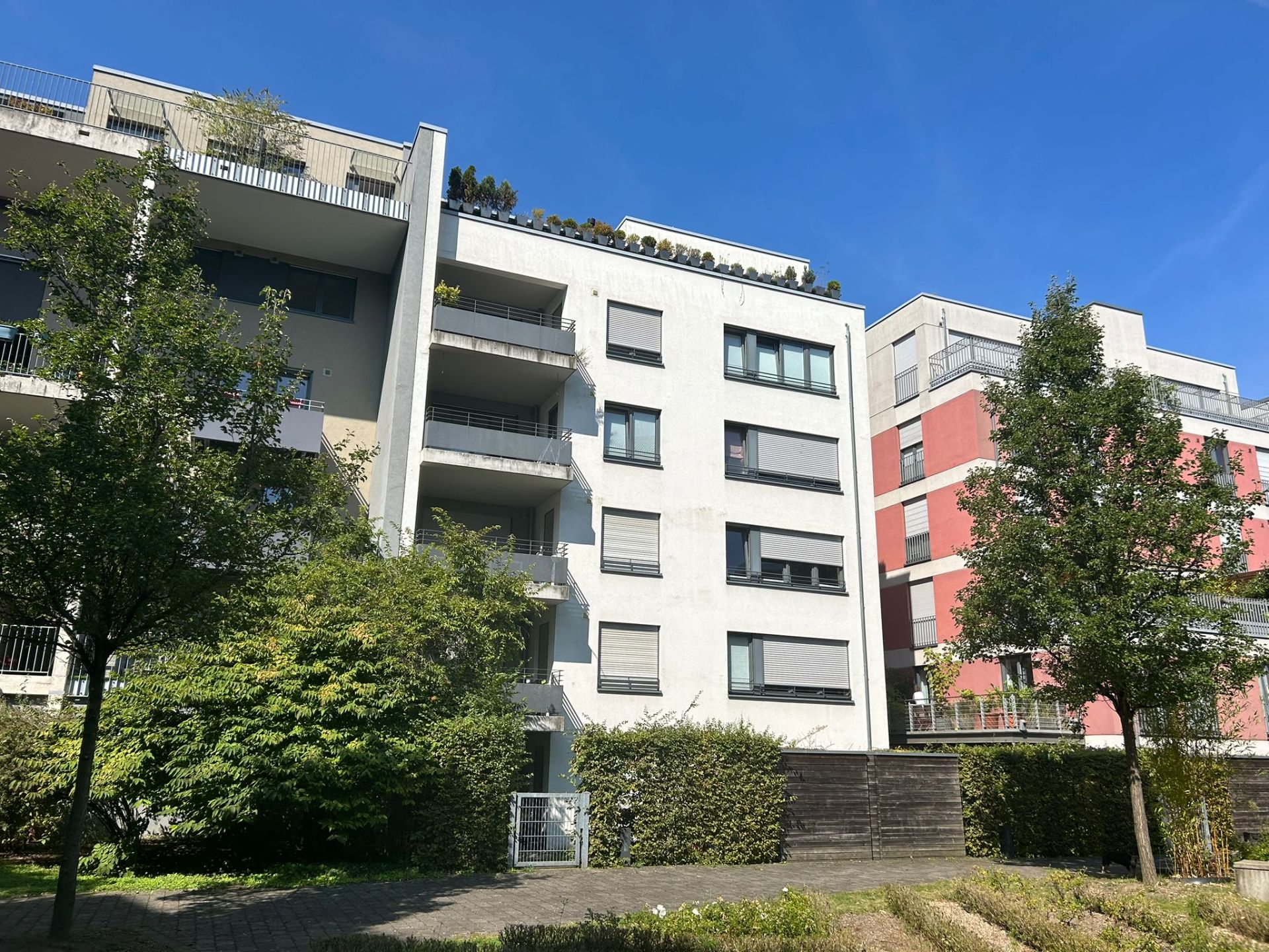 Mehrstöckiges Wohnhaus mit Balkonen, Bäumen und Sträuchern im Vordergrund.
