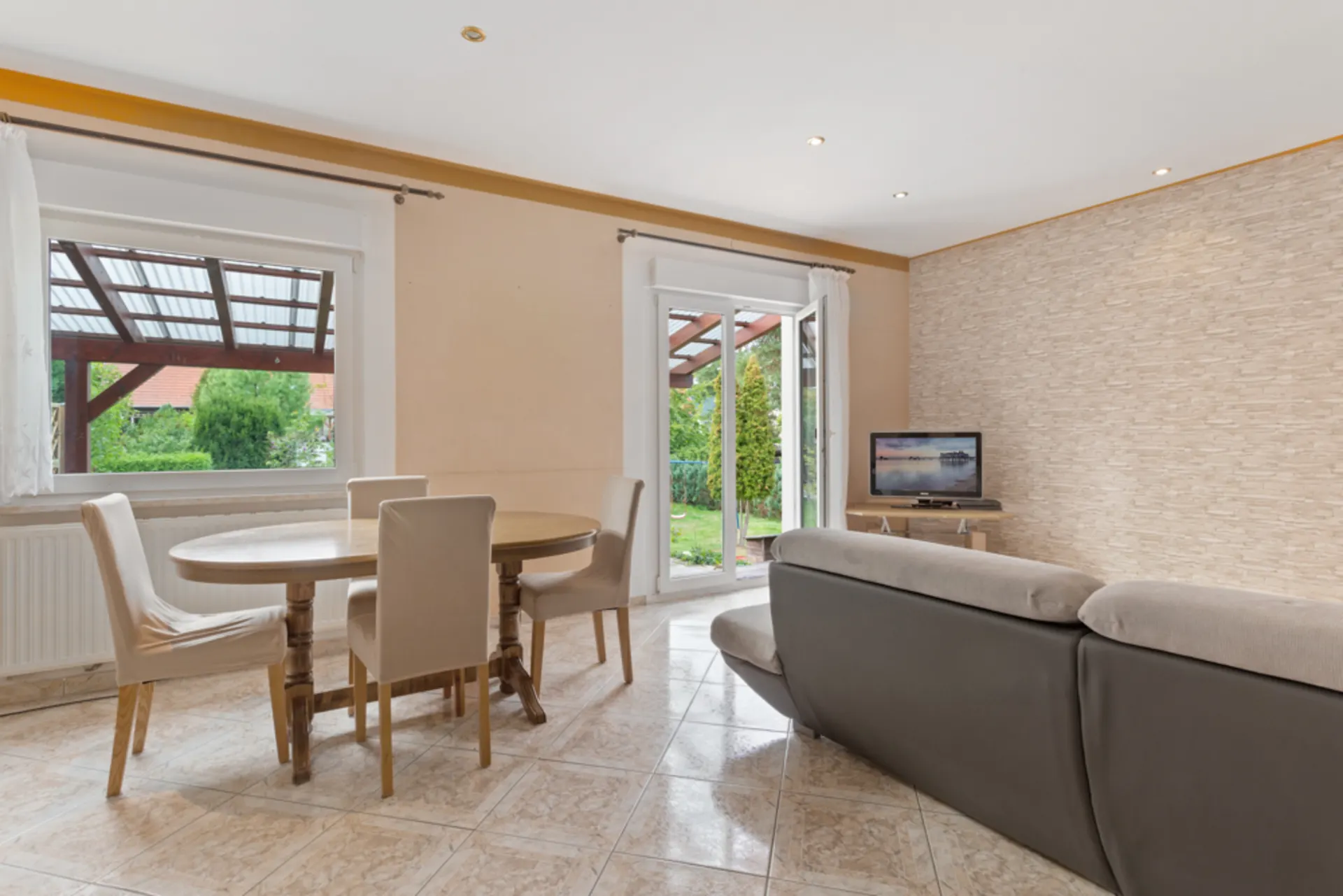 Helles Wohnzimmer mit Esstisch, Stühlen, Sofa und Fernseher, Blick in den Garten.