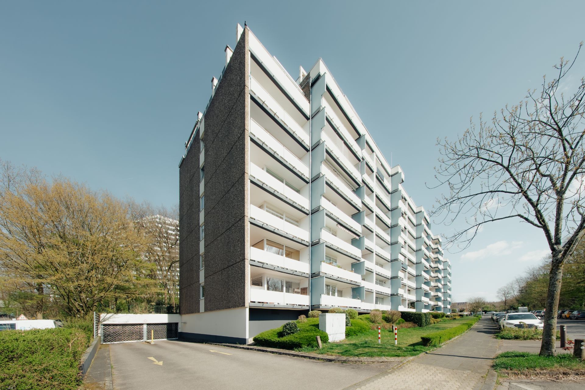 Mehrstöckiges Wohnhaus mit Balkonen, Gehweg und Bäumen im Vordergrund.