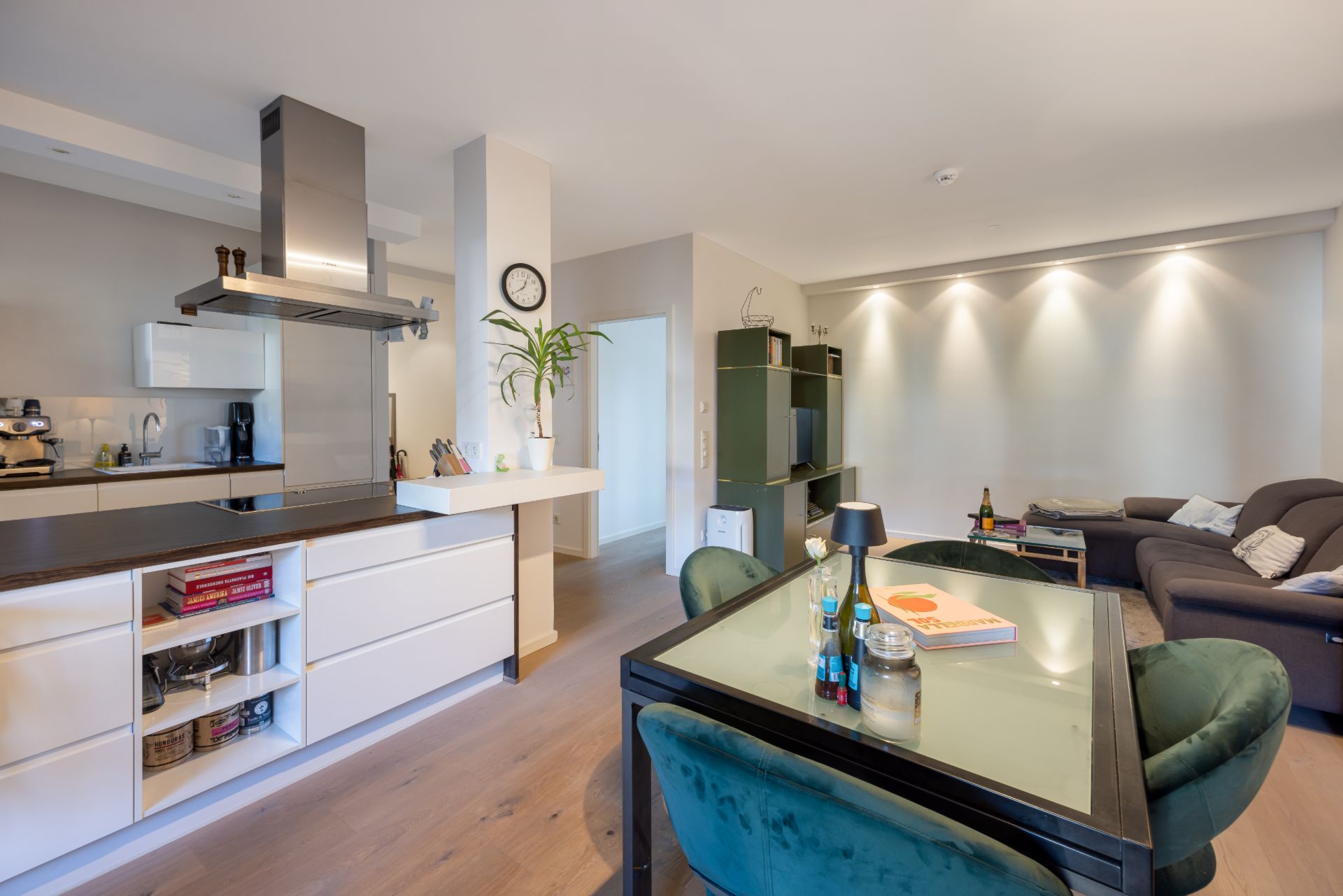 Offene Küche mit Esstisch und angrenzendem Wohnzimmer mit Sofa und Regal, helle Beleuchtung.