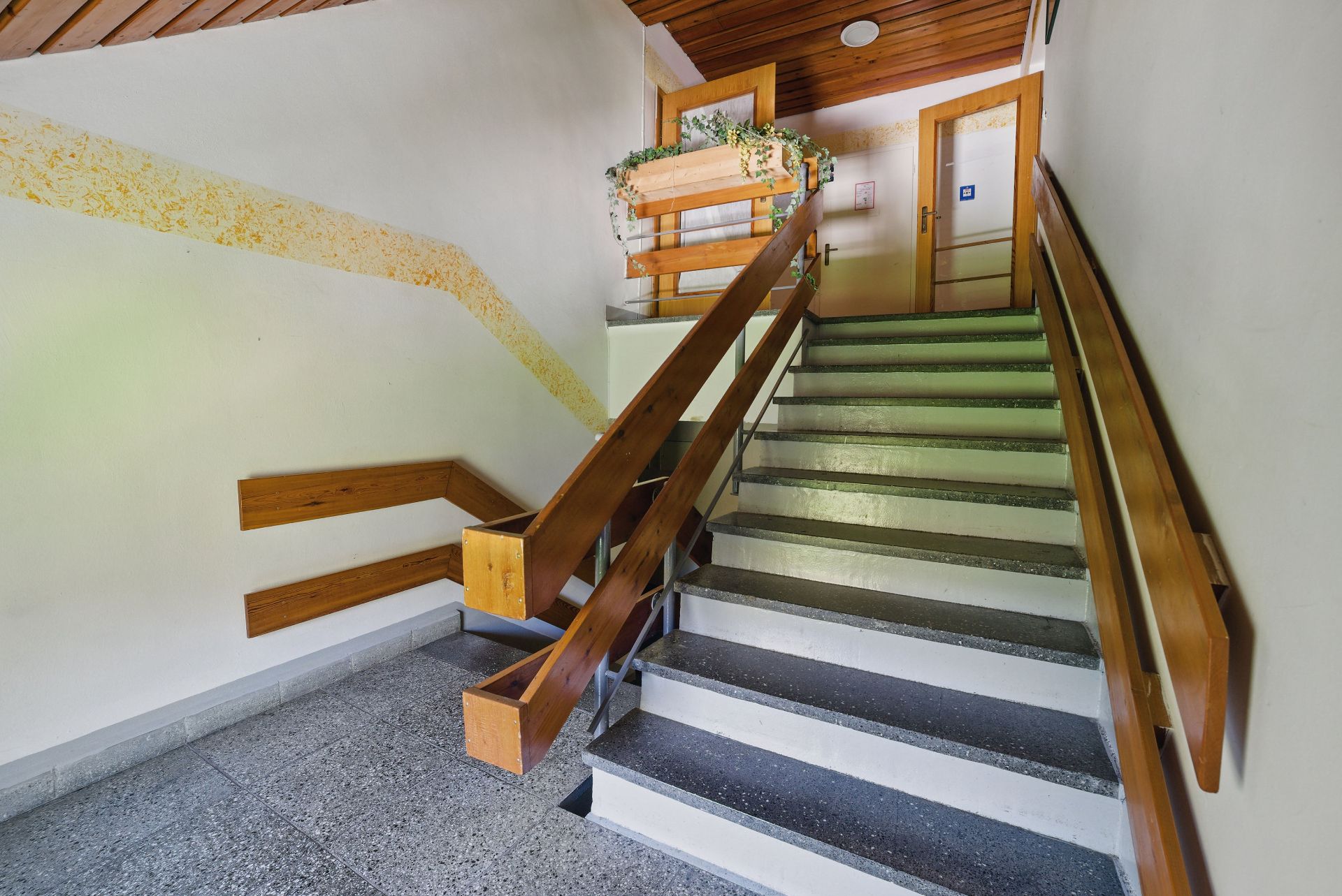Treppenhaus mit Holzgeländer und Pflanzen auf dem Absatz, Türen im oberen Bereich sichtbar.