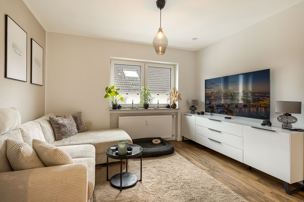 Wohnzimmer mit Ecksofa, Teppich, Couchtisch, Fernseher auf Sideboard und zwei Fenstern.