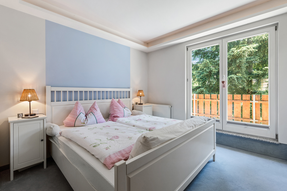 Helles Schlafzimmer mit Doppelbett, Nachttischen und großen Fenstern mit Blick auf Bäume.