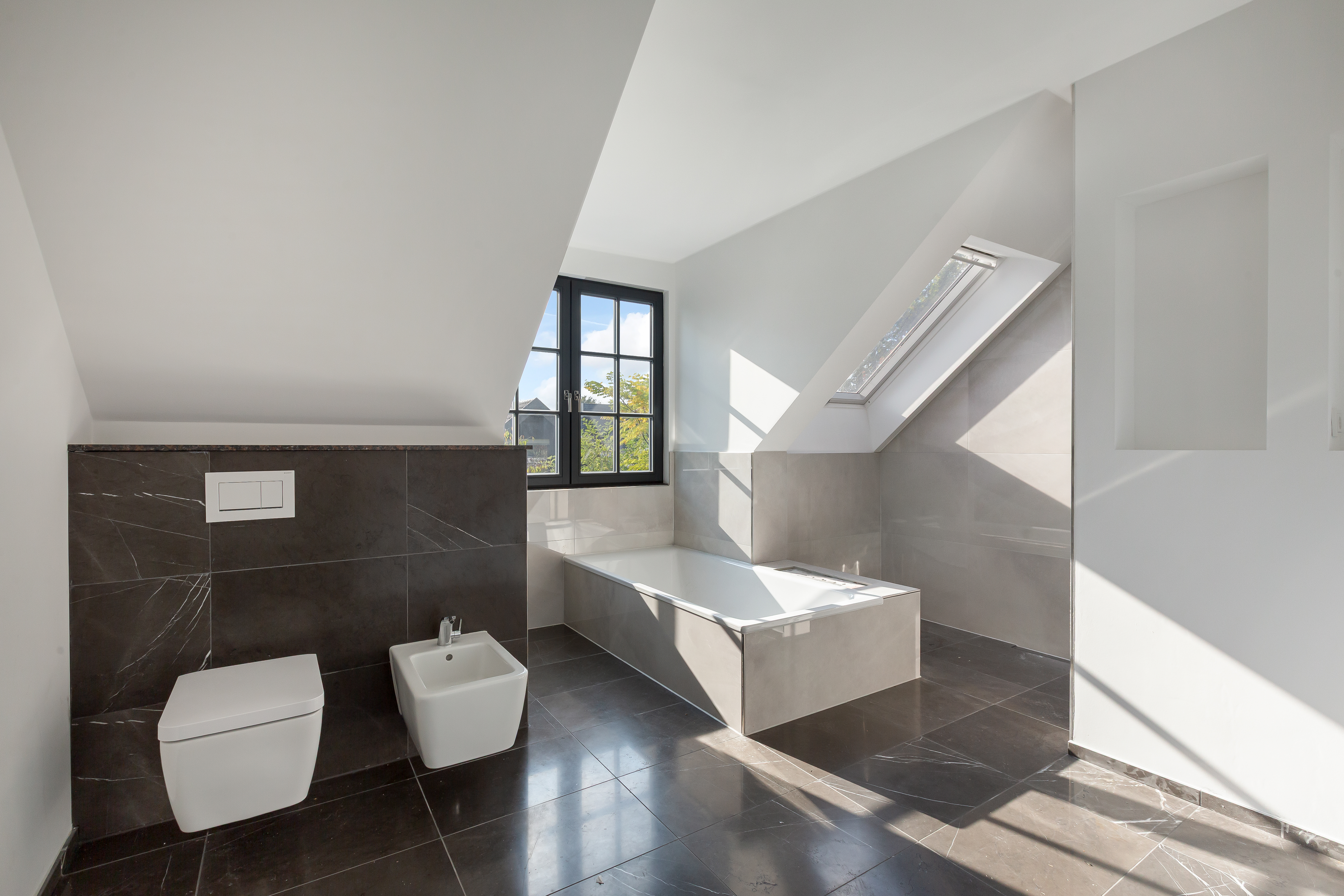 Modernes Badezimmer mit Badewanne, dunklen Fliesen und zwei Fenstern unter einer Dachschräge.