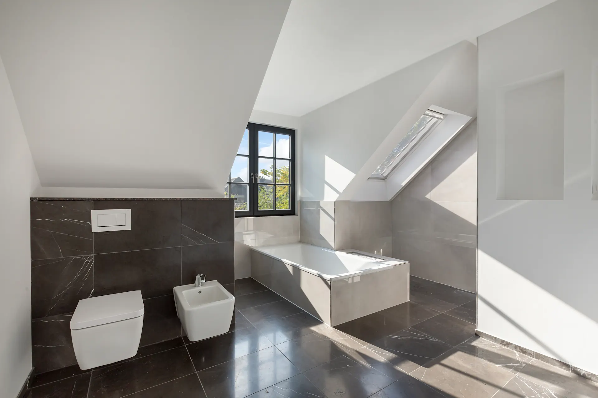 Modernes Badezimmer mit Badewanne, dunklen Fliesen und zwei Fenstern unter einer Dachschräge.