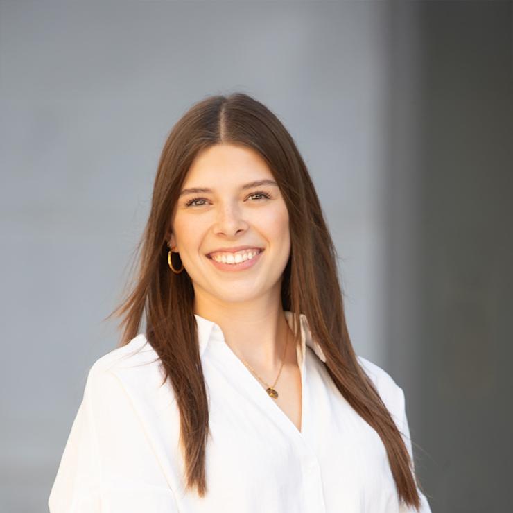 Frau mit langen braunen Haaren und weißer Bluse lächelt vor neutralem Hintergrund.