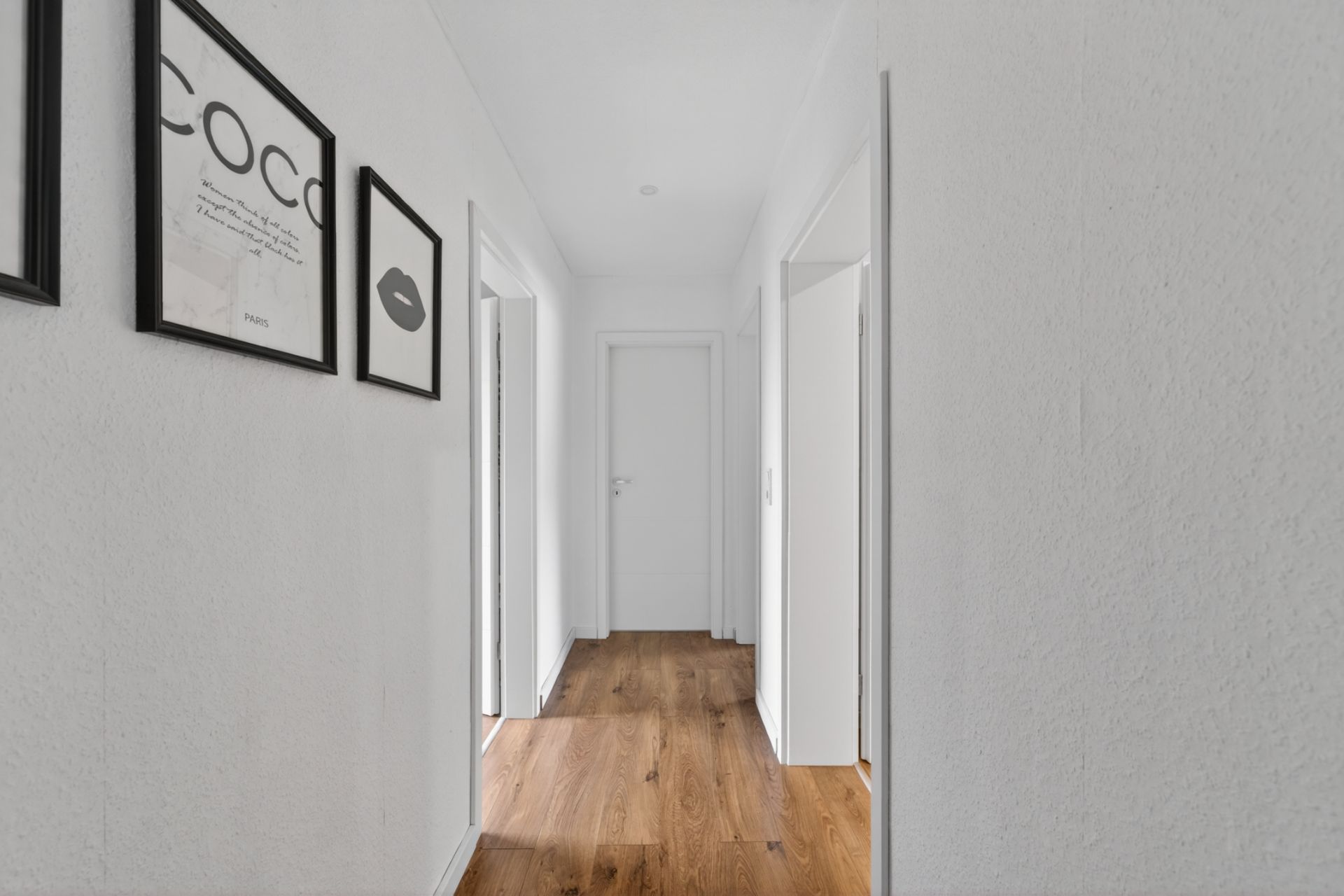 Heller Flur mit Holzfußboden und weißen Wänden, Bilder hängen an der linken Wand.