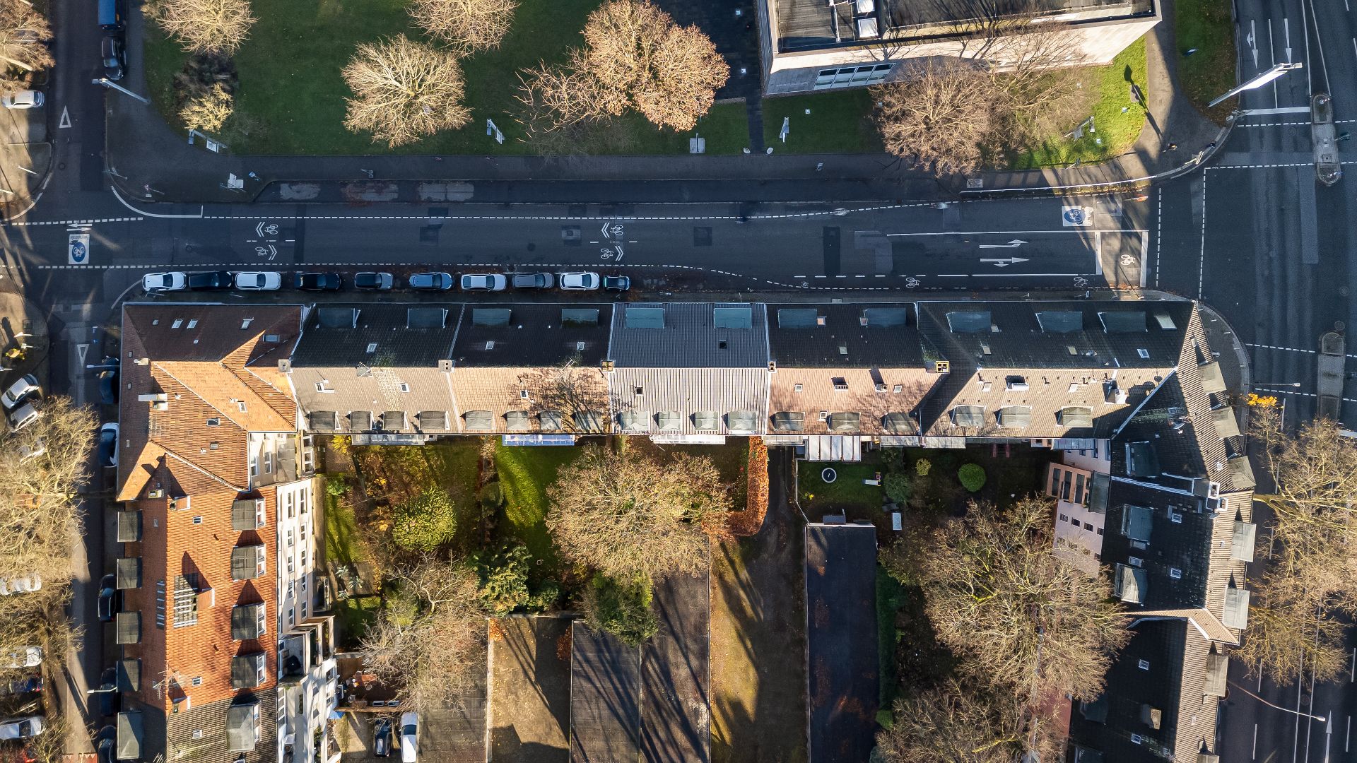 Luftaufnahme eines Wohnblocks mit angrenzenden Gärten und Bäumen sowie geparkten Autos an einer Straße.