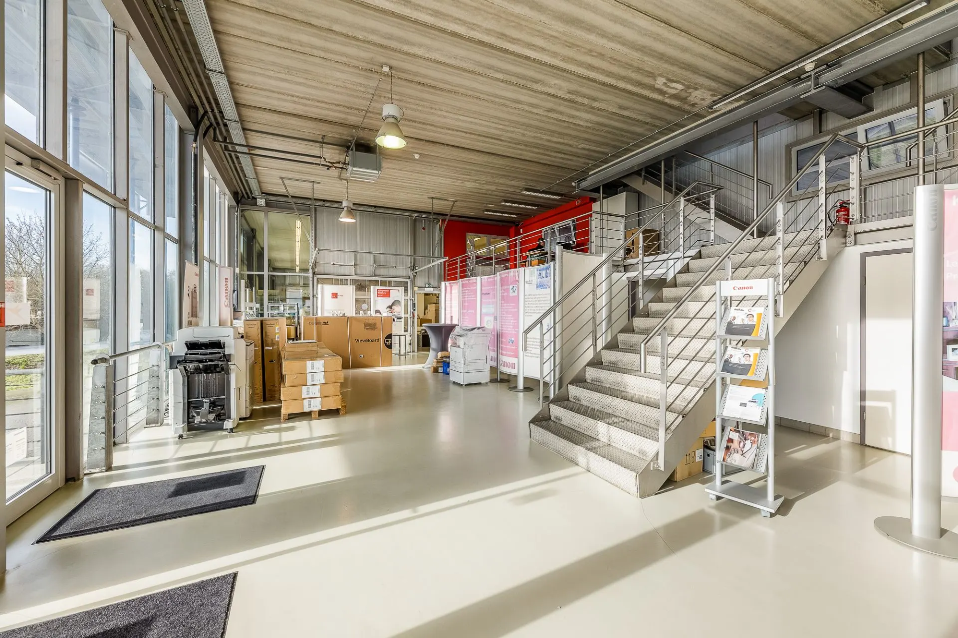 Heller Eingangsbereich mit Treppe, großen Fenstern, Kartons und Prospektständer.