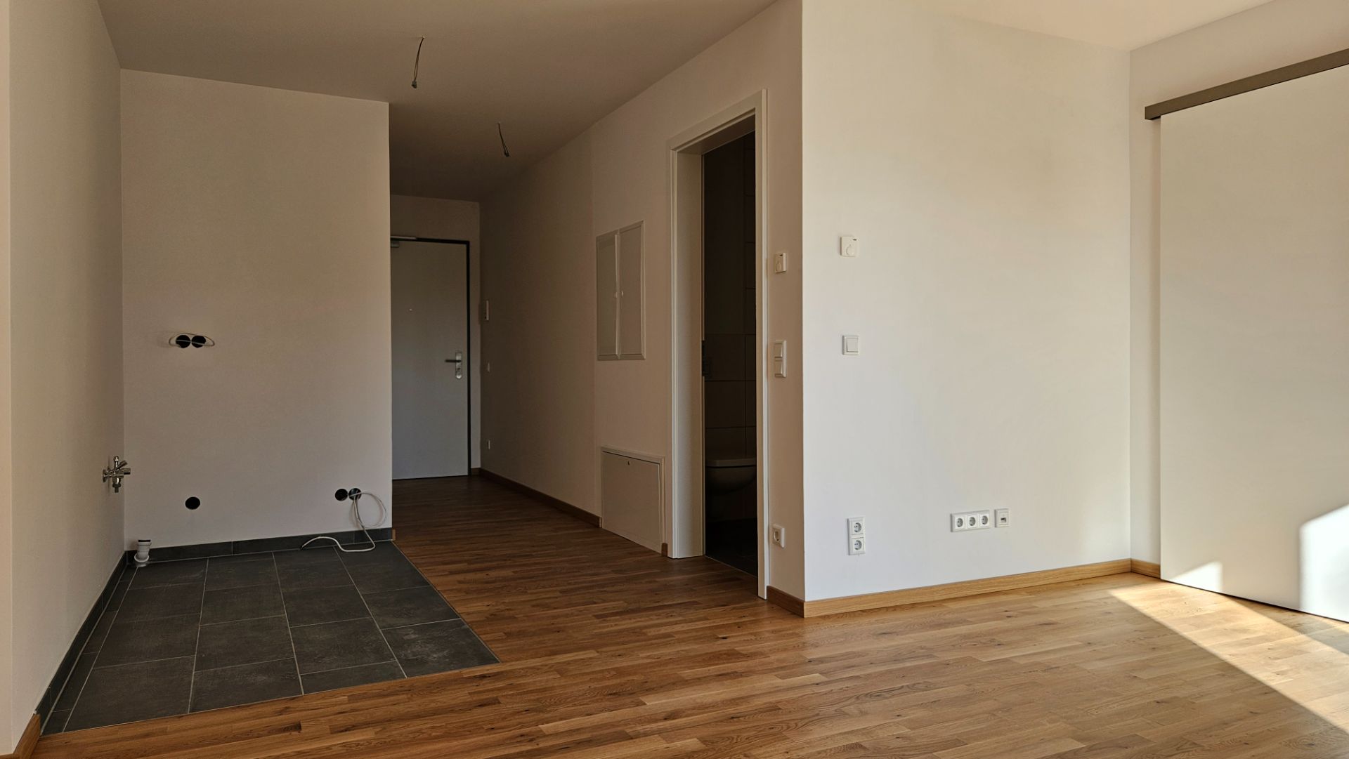 Leerer Raum mit Parkettboden, Fliesenbereich und weiß gestrichenen Wänden, Tageslicht von rechts.
