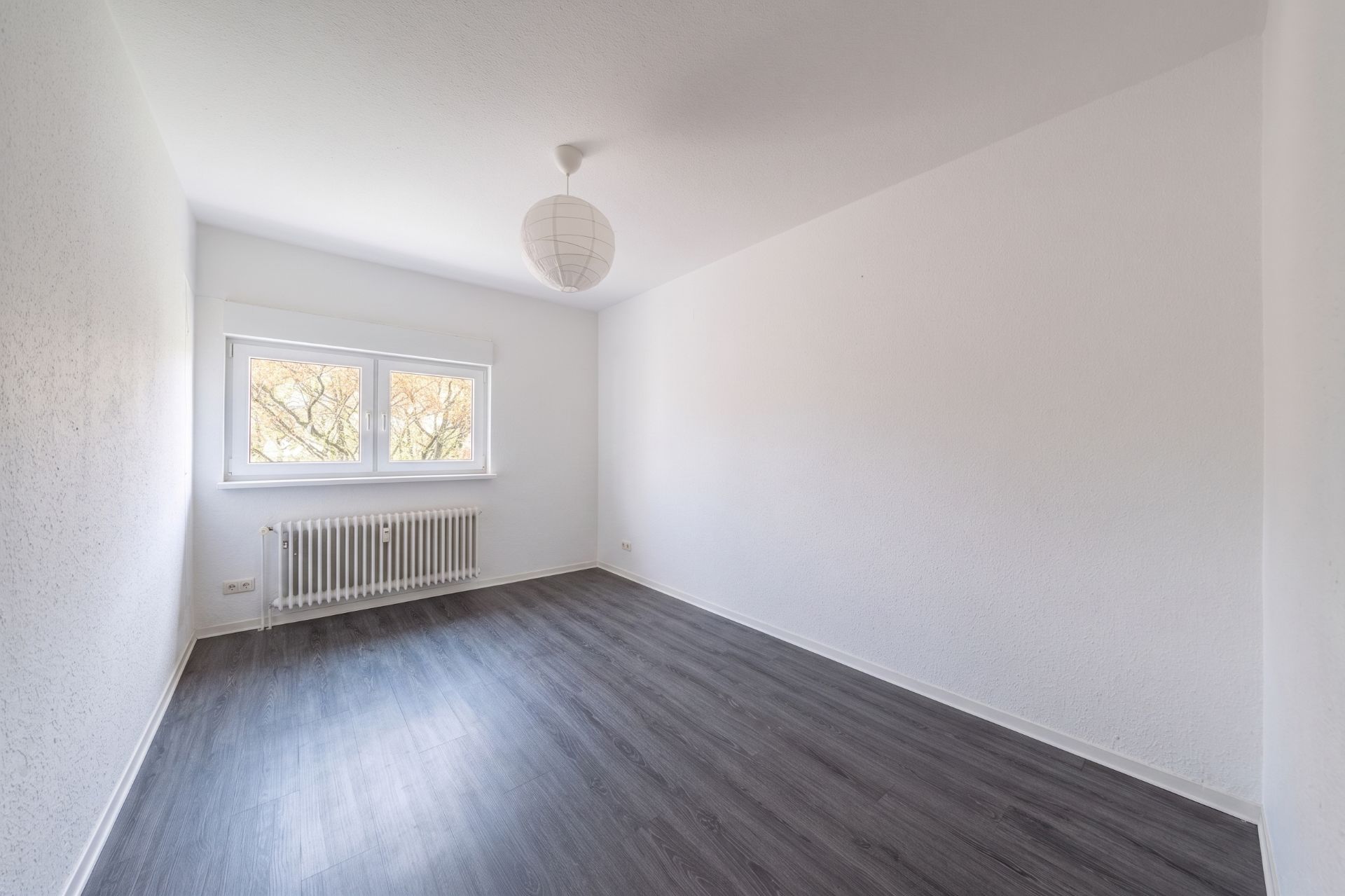 Leeres Zimmer mit weiß gestrichenen Wänden, großem Fenster und dunklem Boden.