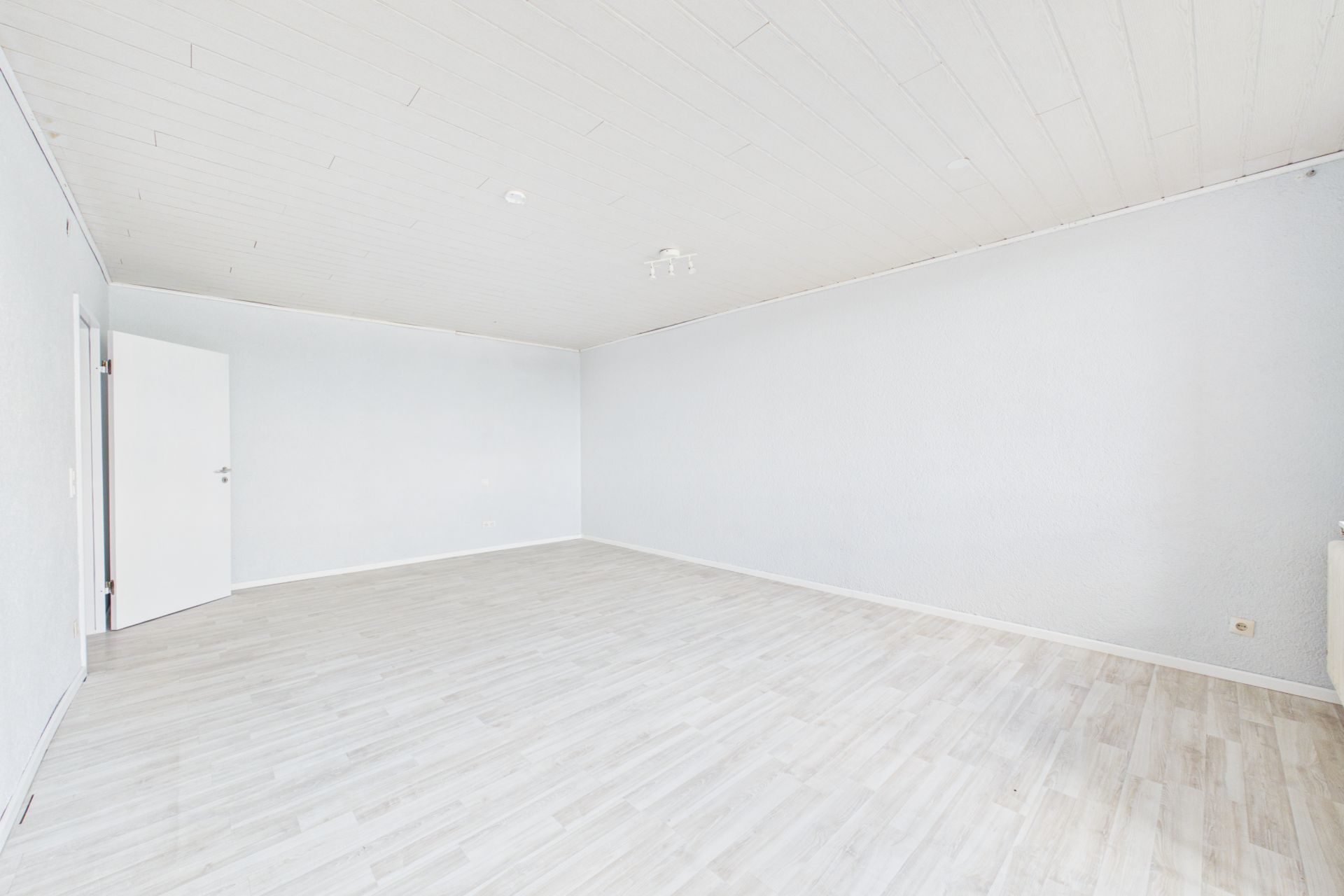 Heller, leerer Raum mit Holzfußboden, weißen Wänden und offener Tür.