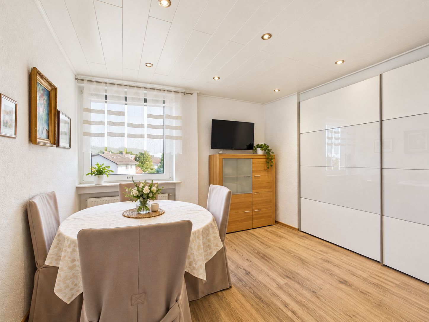 Heller Raum mit Esstisch, vier Stühlen, großem Schrank und Fernseher auf einem Holzschrank.