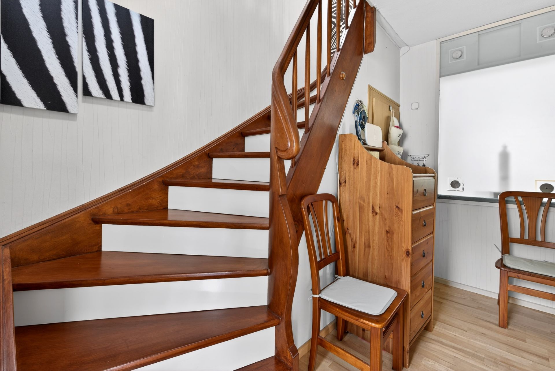 Holztreppe mit zwei Stühlen, kleiner Kommode und Wandbildern in einem hellen Raum.