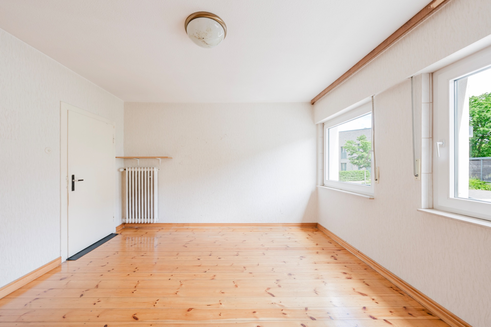 Heller leerer Raum mit Holzboden, zwei Fenstern, Tür und Deckenleuchte.