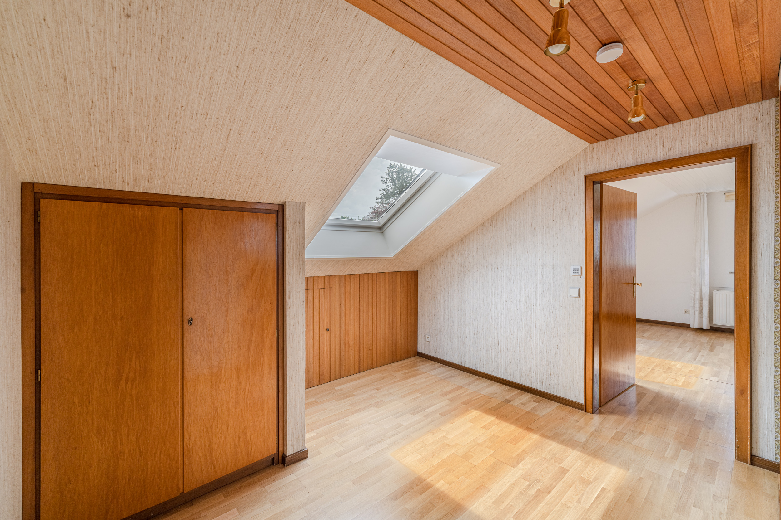 Dachzimmer mit Holzschränken, Dachfenster und Holzboden, offene Tür zum nächsten Raum.
