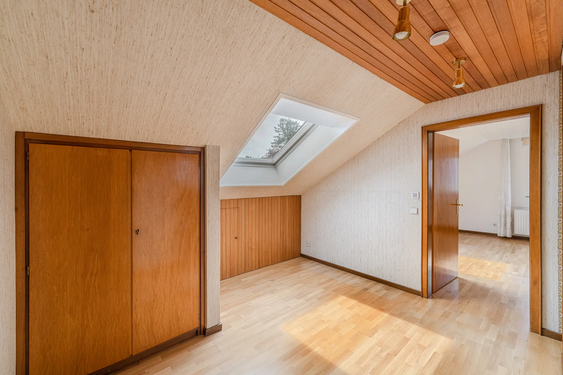 Dachzimmer mit Holzschränken, Dachfenster und Holzboden, offene Tür zum nächsten Raum.