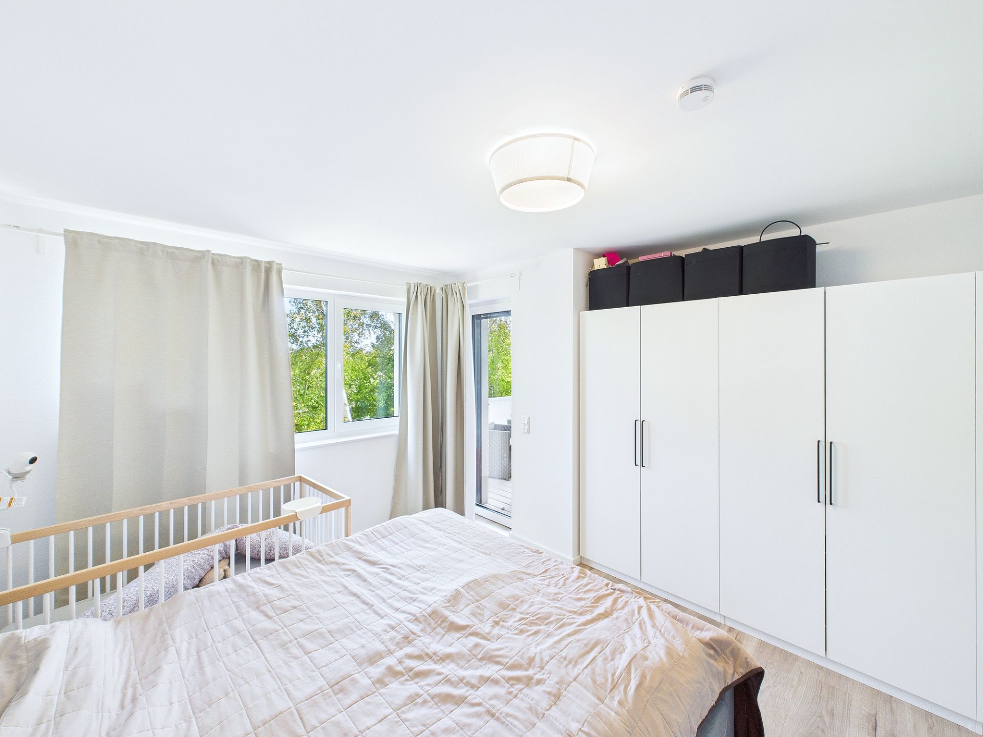 Helles Schlafzimmer mit Doppelbett, Babybett, weißen Vorhängen und großem weißen Kleiderschrank.
