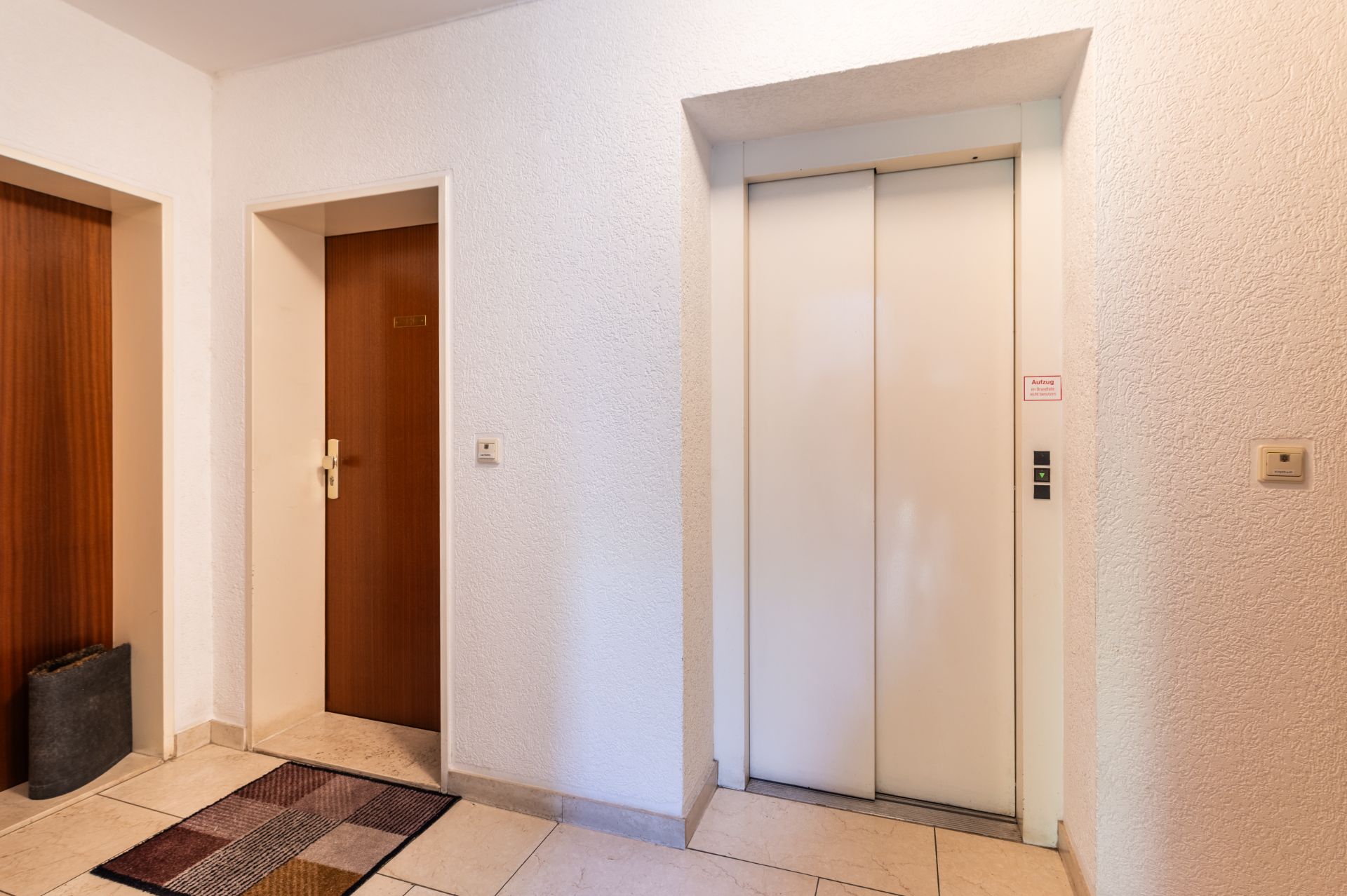 Flur mit Aufzug, zwei Wohnungstüren und einem Teppich auf dem Boden.