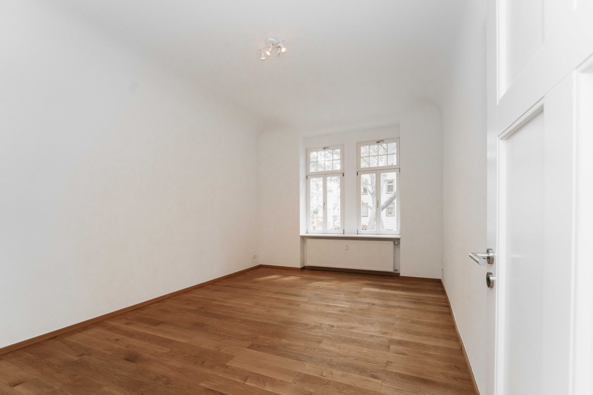 Helles, leeres Zimmer mit Holzfußboden und großem Fenster.
