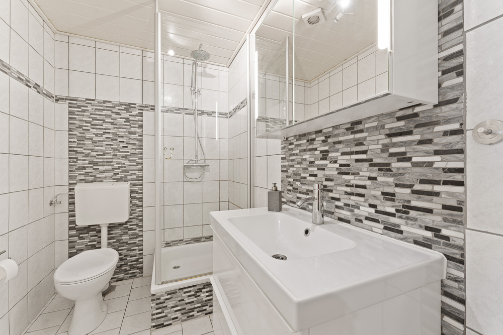 Modernes Bad mit Dusche, WC und Waschbecken, weiße Fliesen und gemusterte Mosaikwand.