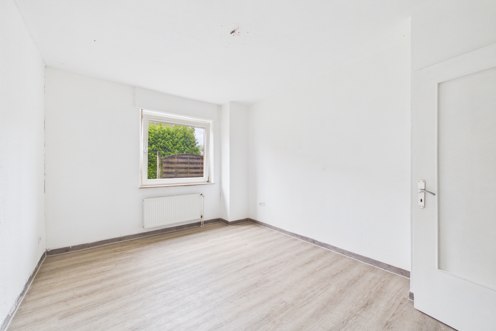 Helles, leeres Zimmer mit weißer Wand, großem Fenster und hellem Holzboden.