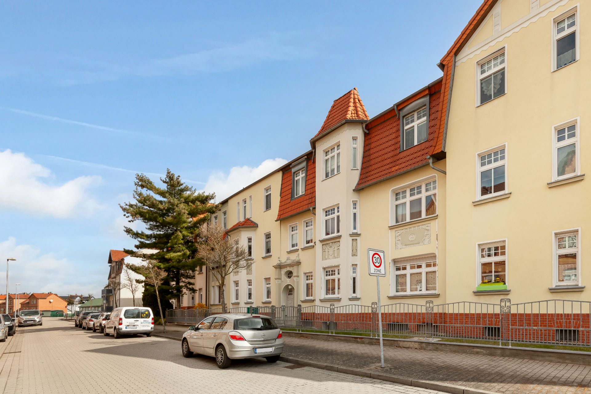 Mehrfamilienhaus mit roten Dächern an einer Wohnstraße, Autos sind am Straßenrand geparkt.