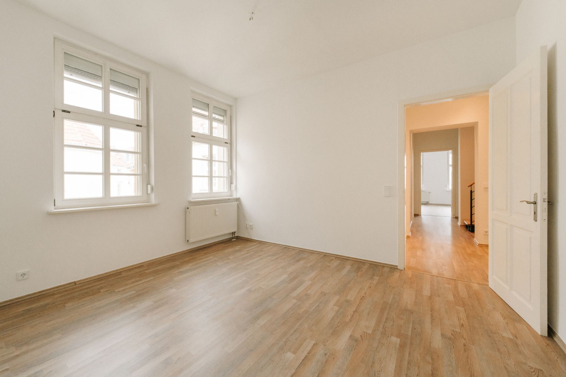 Heller, leerer Raum mit Holzboden, weißen Wänden und zwei Fenstern.