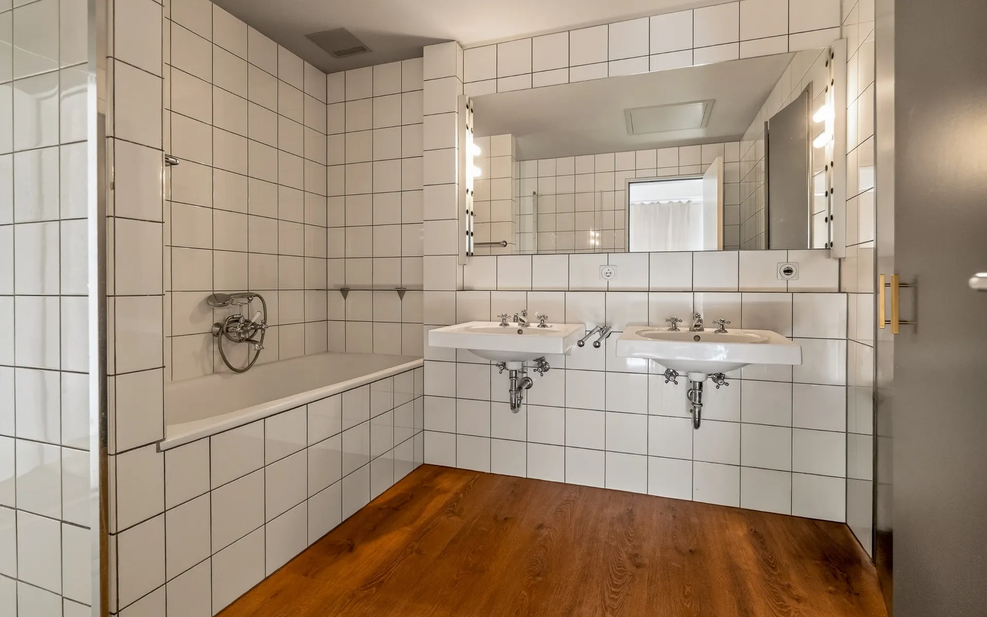 Helles Badezimmer mit zwei Waschbecken, Badewanne, weiß gefliest und Holzboden.