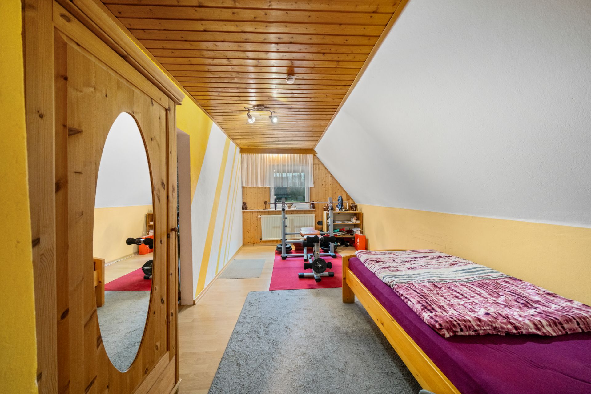 Zimmer mit Holzdecke, Einzelbett, Kleiderschrank mit Spiegel und Fitnessgeräten.