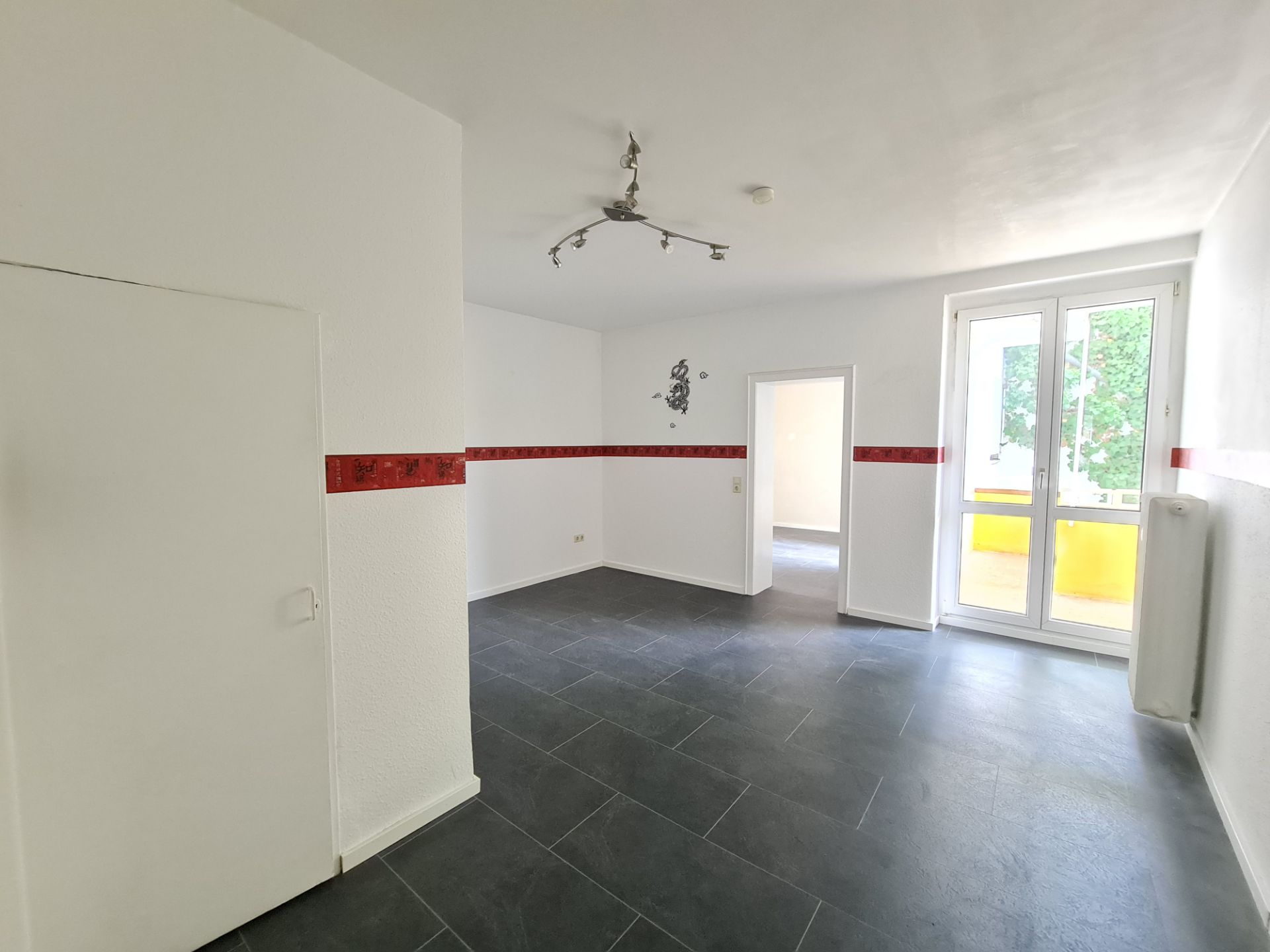 Helles Zimmer mit grauem Fliesenboden, rotem Wandstreifen und Fenstertür zum Balkon.