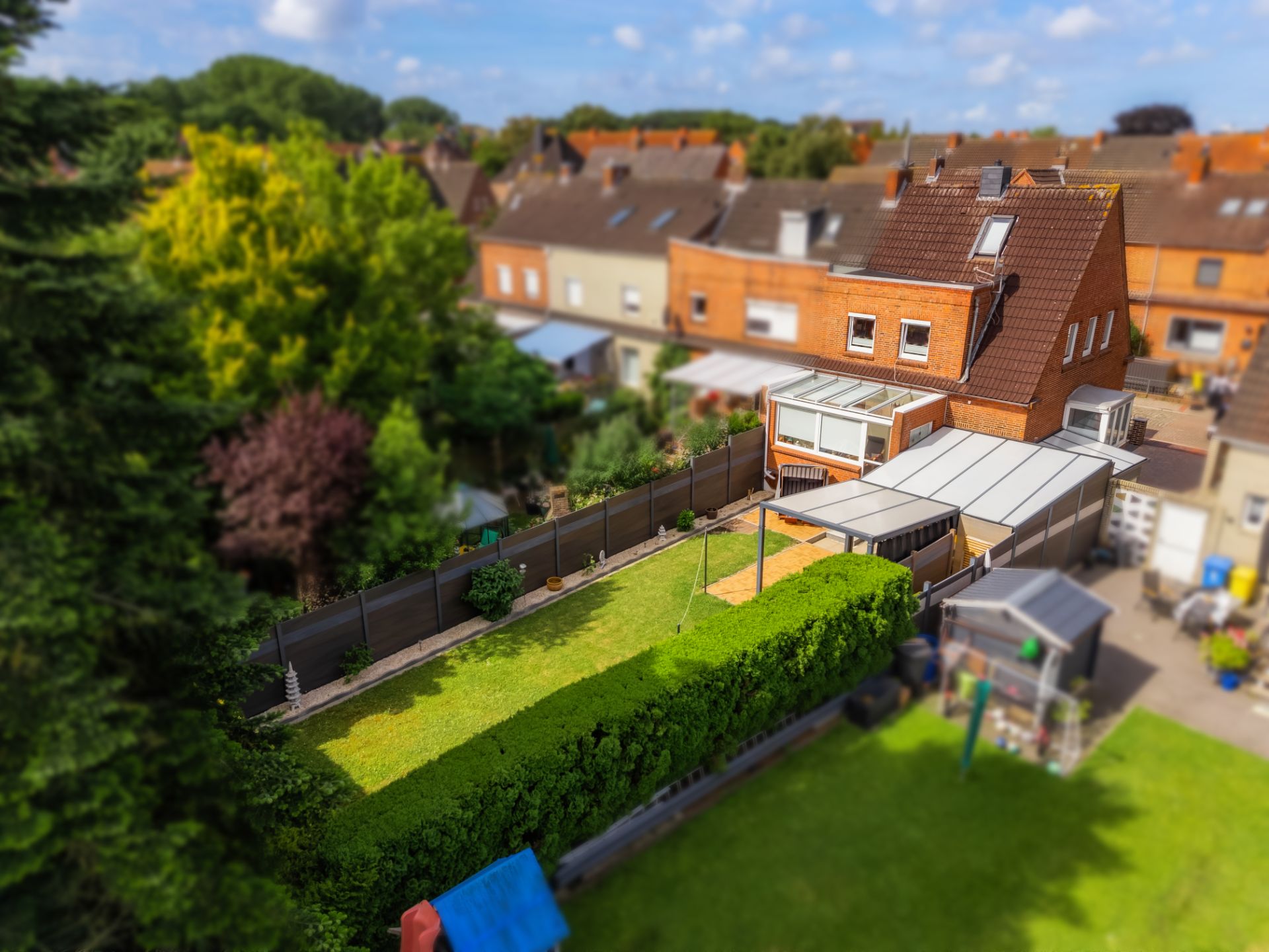 Backsteinhaus mit Garten, Rasenfläche, Schuppen und überdachter Terrasse, umgeben von Hecken.