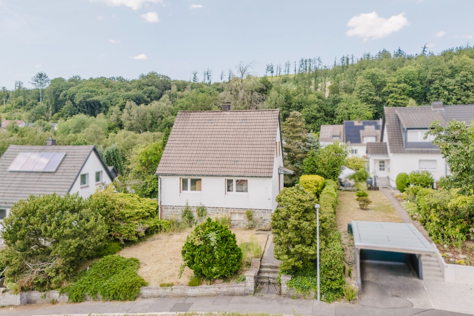 Einfamilienhaus mit Garten und Carport, umgeben von Bäumen und Nachbarhäusern.