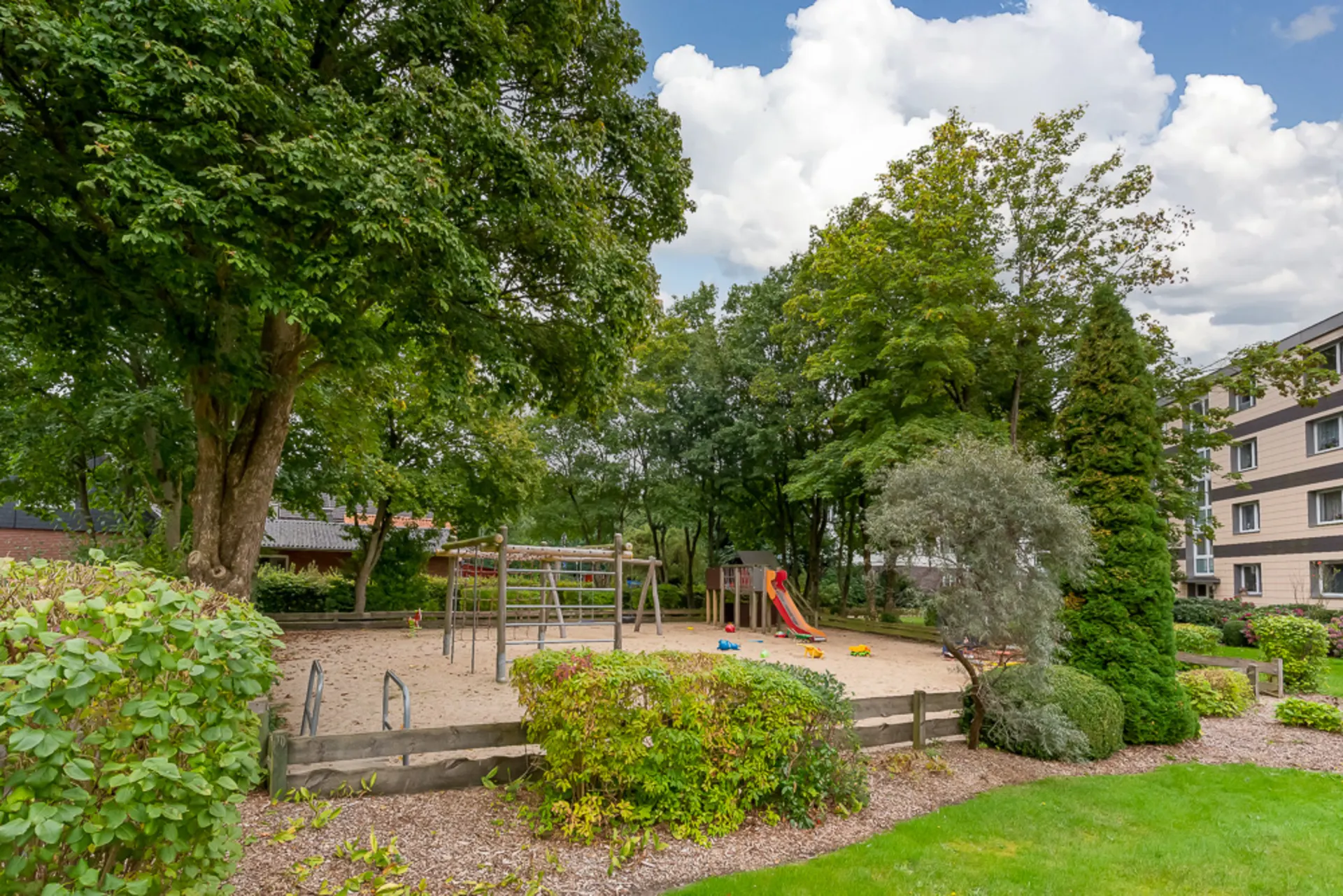 Spielplatz mit Klettergerüst und Rutsche, umgeben von Bäumen und Wohngebäuden.