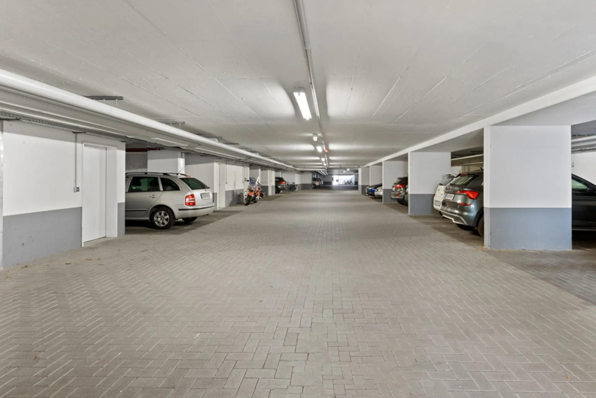 Helle Tiefgarage mit parkenden Autos und Motorrädern, weiße Wände und helle Beleuchtung.