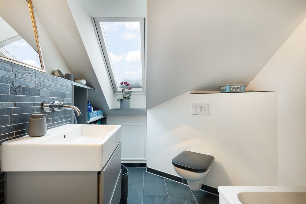 Modernes Bad mit Dachfenster, Waschbecken, Wandtoilette und grauen Fliesen.