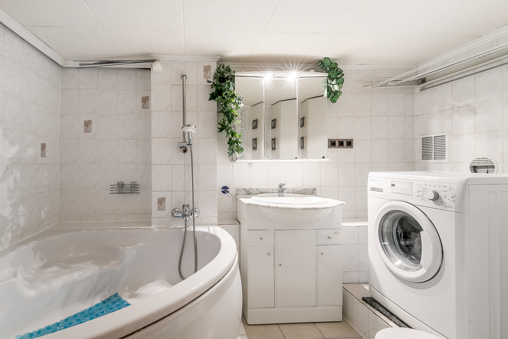 Helles Badezimmer mit Eckbadewanne, Waschmaschine, Waschtisch und Spiegel mit Pflanzen.