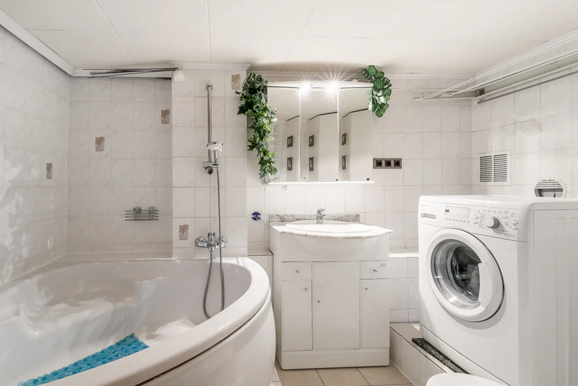 Helles Badezimmer mit Eckbadewanne, Waschmaschine, Waschtisch und Spiegel mit Pflanzen.