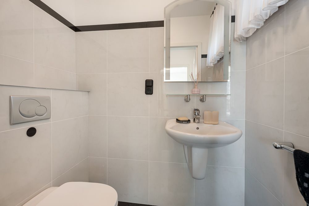 Kleines Badezimmer mit Wandtoilette, Waschbecken, Spiegel und Fenster mit Vorhang.
