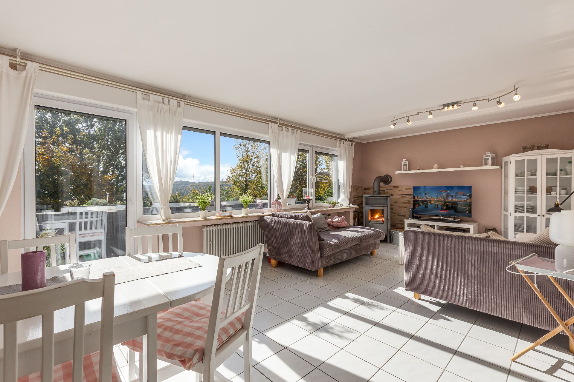 Helles Wohnzimmer mit Sofas, Esstisch, Kaminofen, TV und großen Fenstern mit Blick ins Grüne.