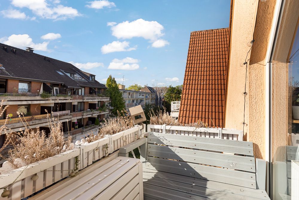 Balkon mit Holzbank und Pflanzen, Blick auf benachbarte Wohnhäuser und blauen Himmel.