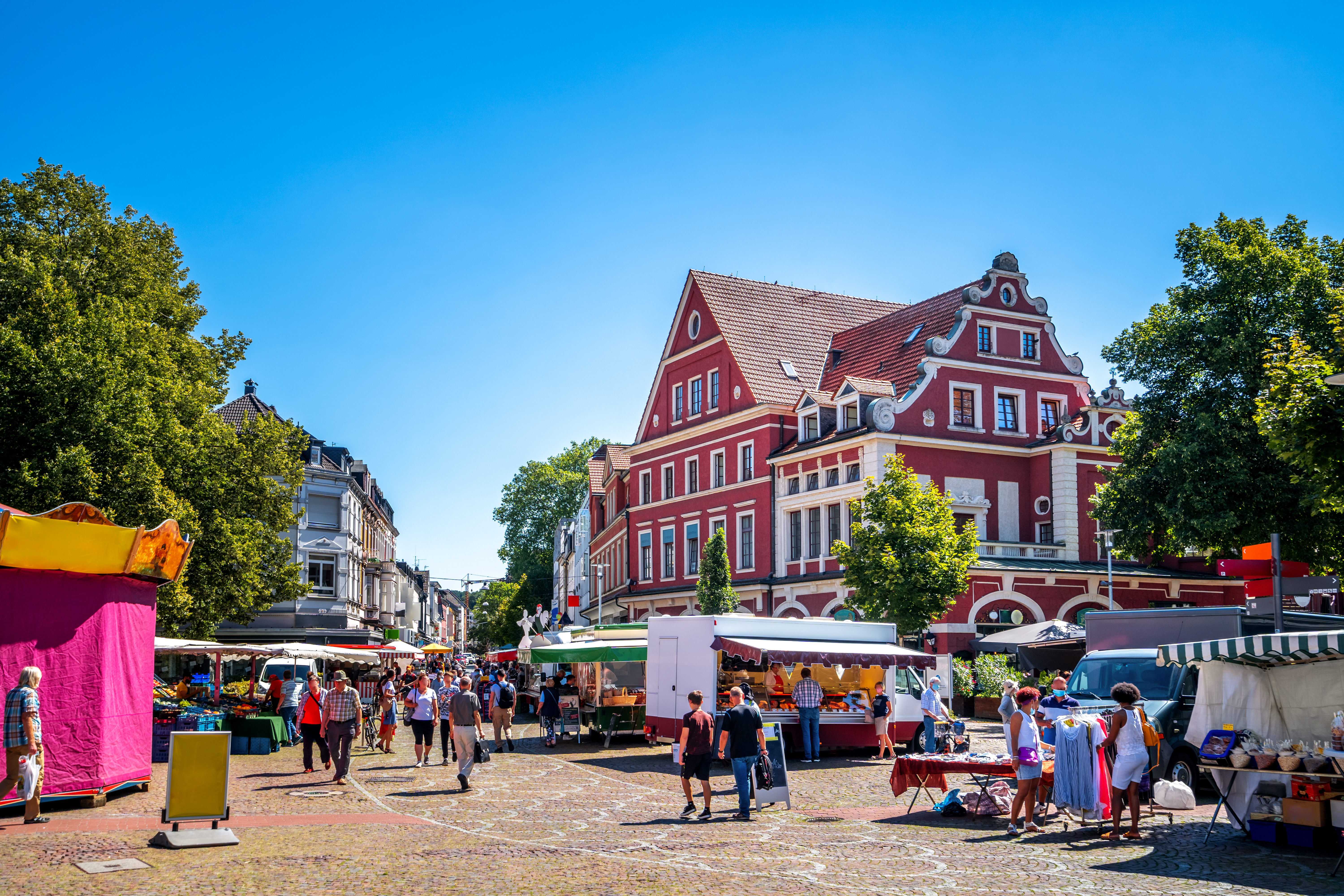 Menschen schlendern auf einem belebten Marktplatz mit Ständen und einem roten Gebäude im Hintergrund.