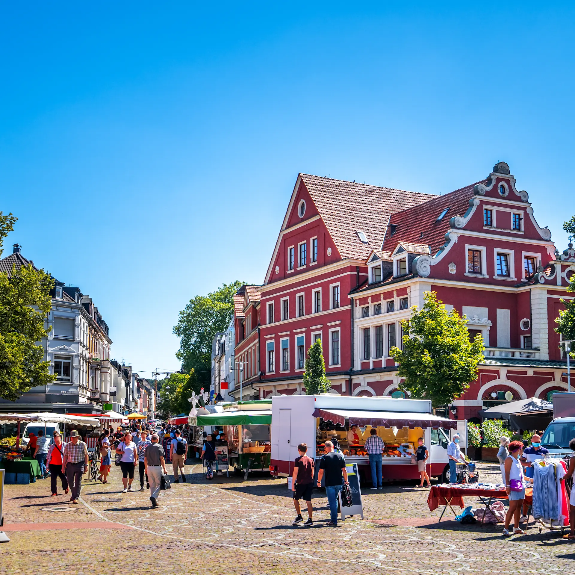 Menschen schlendern auf einem belebten Marktplatz mit Ständen und einem roten Gebäude im Hintergrund.