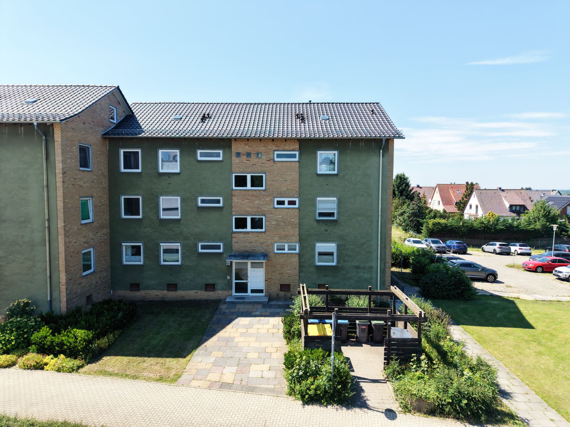 Mehrfamilienhaus mit grünem Putz, Vorgarten und Parkplatz im Hintergrund bei Tageslicht.