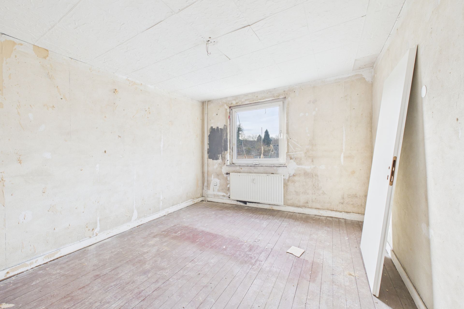 Leerer Raum mit abgenutztem Holzboden, offenen Wänden, Fenster und angelehnter Tür.