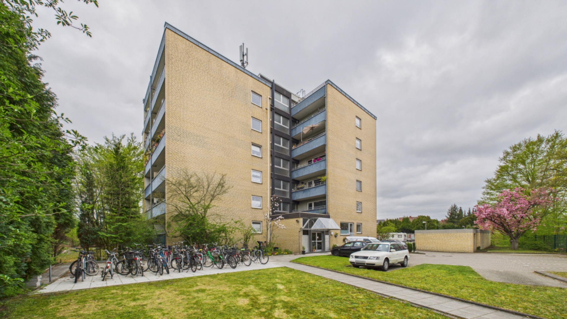 Mehrstöckiges Wohnhaus mit Balkonen, Fahrrädern und geparkten Autos vor dem Eingang.