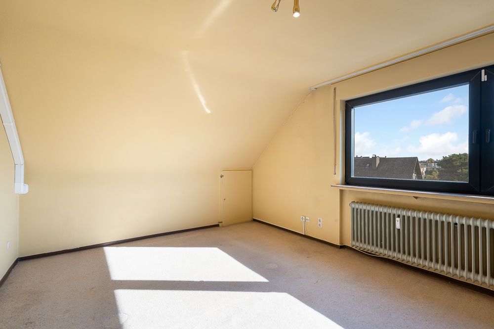 Leeres Zimmer mit Dachschräge, großem Fenster und Heizkörper unter dem Fenster.