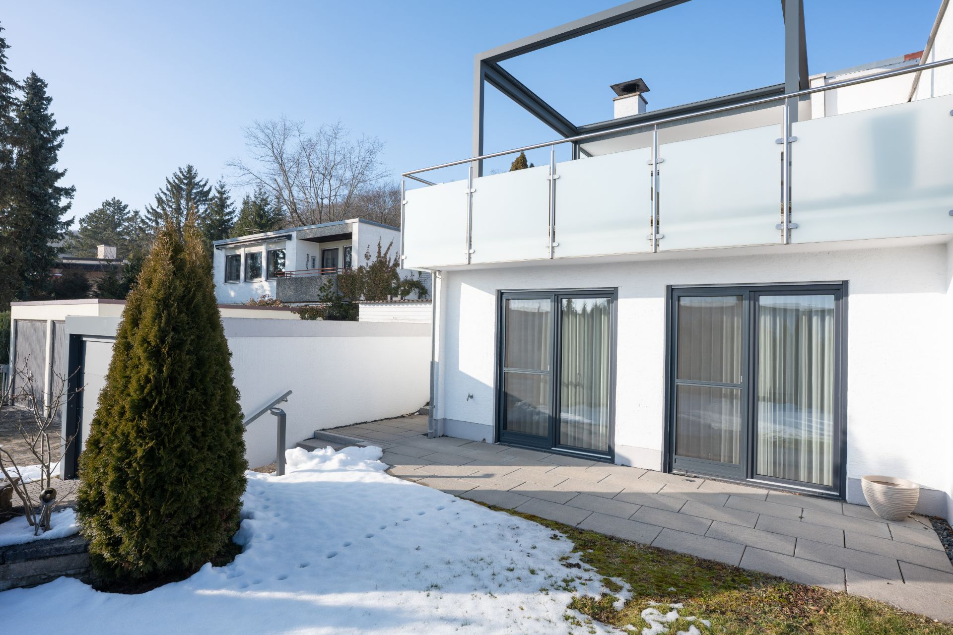 Moderne Terrasse mit Glastüren, etwas Schnee und einer grünen Hecke vor einem Wohnhaus.