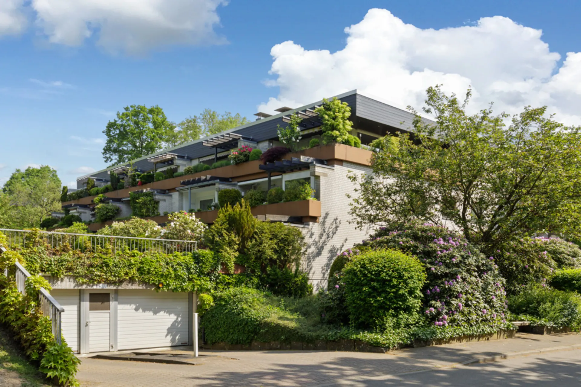 Mehrfamilienhaus mit mehreren Balkonen und vielen Pflanzen, umgeben von Bäumen und Sträuchern.
