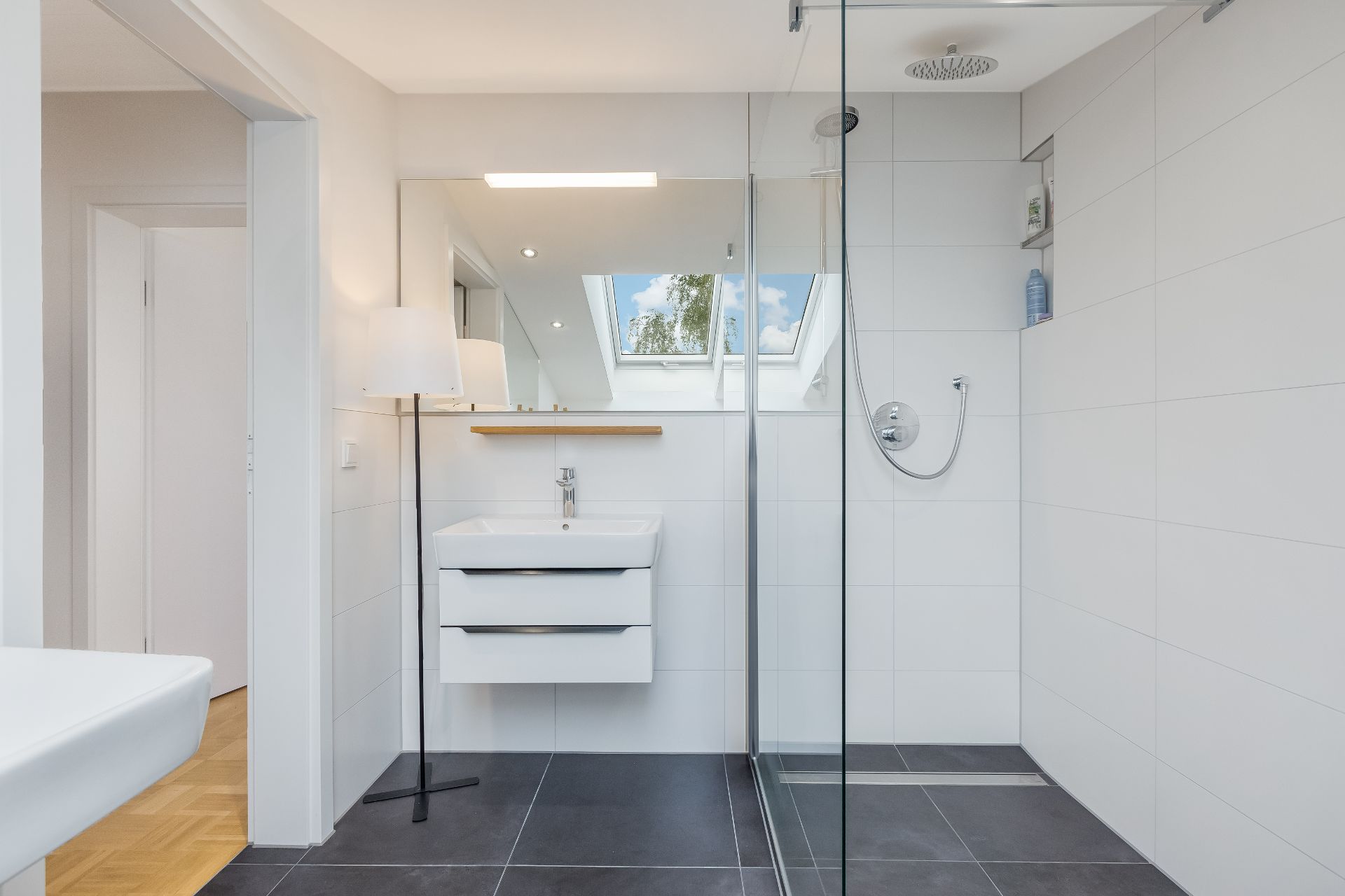 Modernes Badezimmer mit Dusche, Waschbecken und großem Spiegel unter einem Dachfenster.